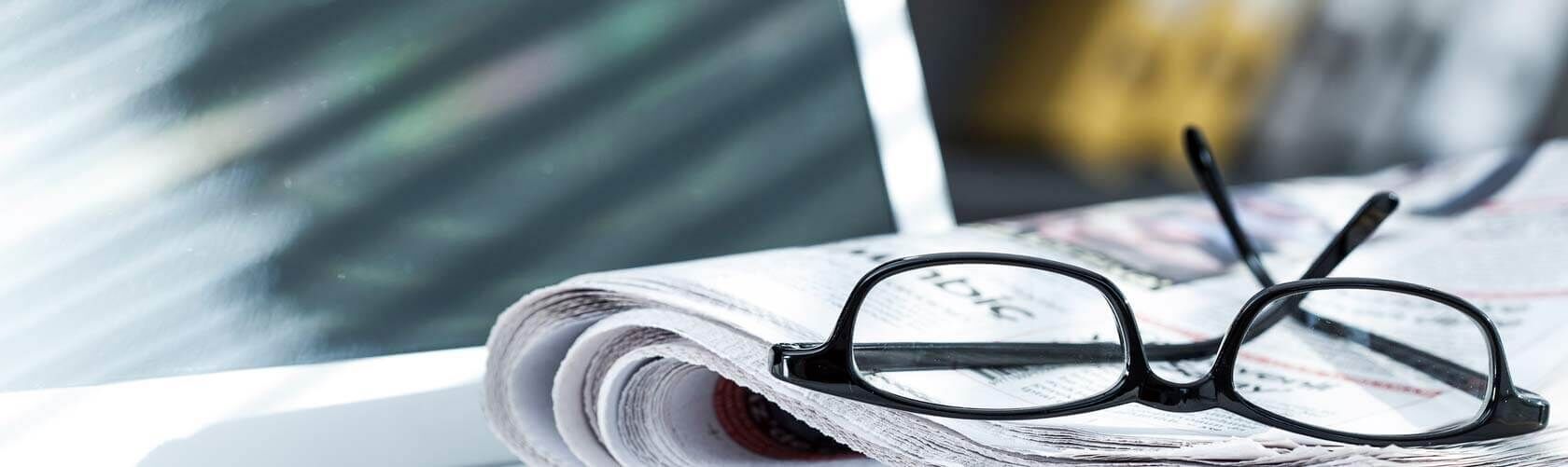 Newsletter Abonnement Eine Brille liegt auf einem Stapel Zeitungen, beleuchtet von sanftem Tageslicht durch ein Fenster.